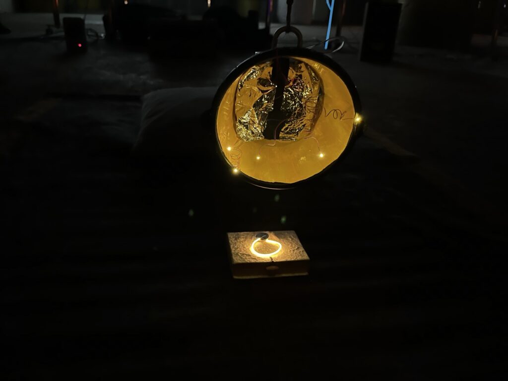 Close view of a small speaker wrapped in reflective plastic foil in the center of a yellow cone illuminated with tiny white lights. It's all suspended from a rope, against a dark background.