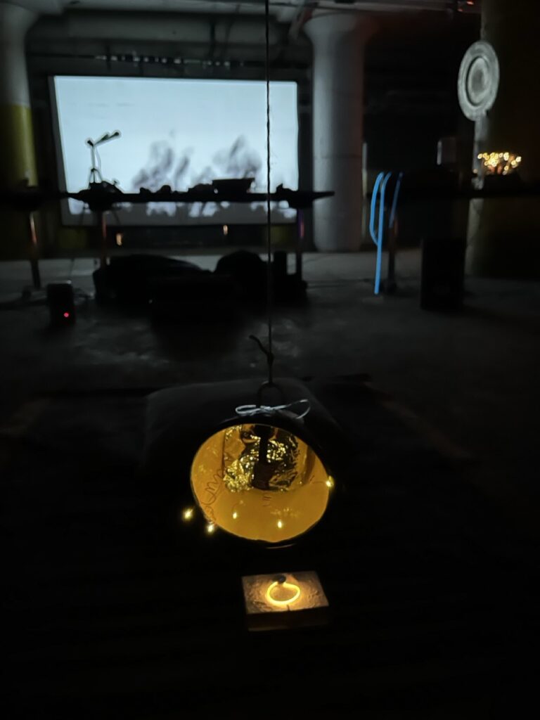 A small speaker wrapped in reflective plastic foil in the center of a yellow cone illuminated with tiny white lights. It's all suspended from a rope. In the background is a performance space with tables cluttered with instruments, all silhouetted against a projection screen