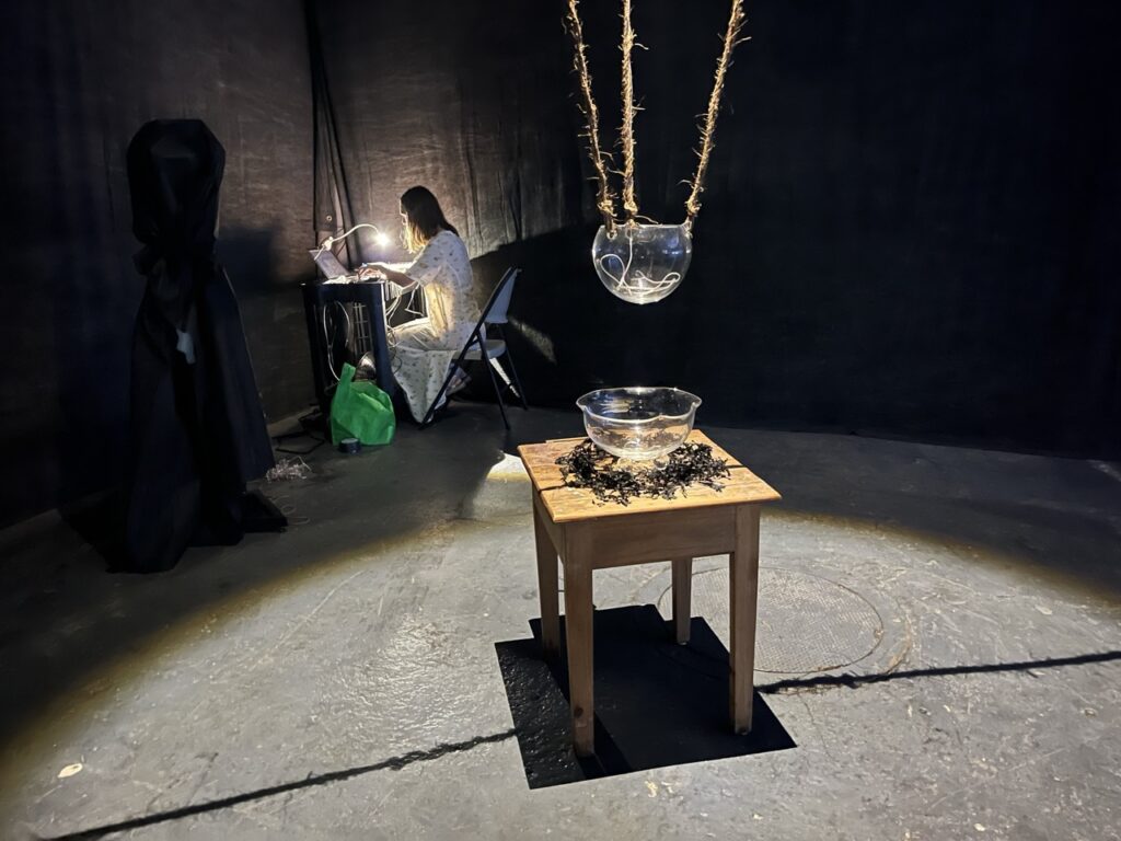 Close view of spotlit glass vessels, one suspended from above by ropes and the other on a small wooden table below. In the background, Anne Hollænder works on a computer on music for the installation. The black-shrouded speaker is visible just outside of the spotlight.