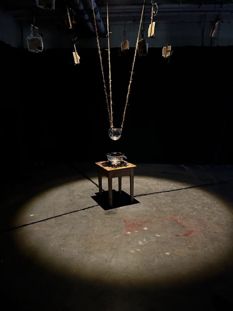 a glass vessel hangs from the ceiling in a white circle of light against a stark black background. Another vessel lies directly below it on a small wooden table.