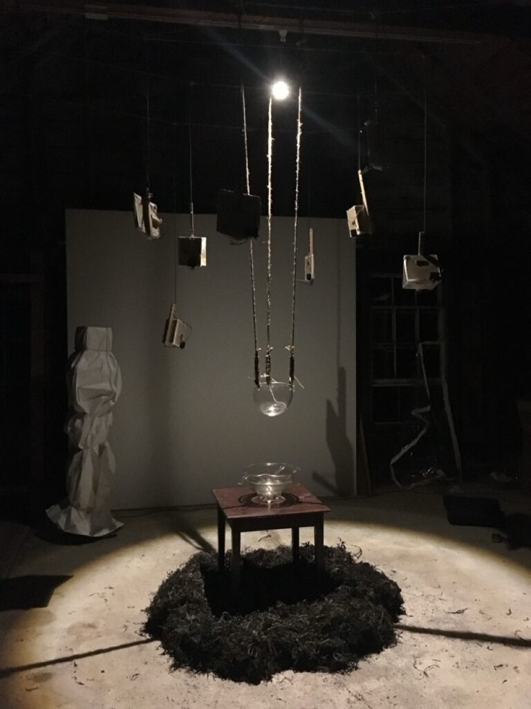 In a dark room, an overhead spotlight illuminates a circle on the floor. Small instruments and a glass vessel hang from the ceiling; another vessel is on a small table in the middle of a pile of seaweed. In the background outside of the circle of light, a speaker on a stand is wrapped in white fabric, looking a bit like a ghostly human figure.