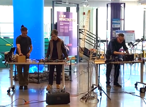 Three people stand behind tables, manipulating objects and instruments.